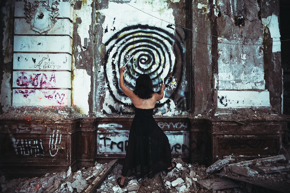A female drummer juggler in a sheer, black dance dress playing the graffiti-ridden wall of a dilapidated auditorium with drumsticks.