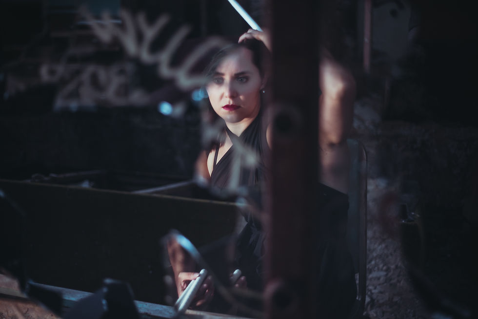 A female drummer juggler wearing a sheer, black dance dress seeing her reflection in the shattered mirror of a vandalized makeup table.