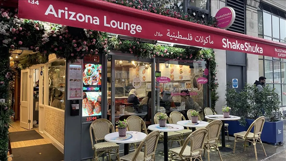 Outdoor view of Arizona Lounge with red awning. Tables and chairs outside, greenery decor, visible signage with colorful menus. Busy atmosphere.