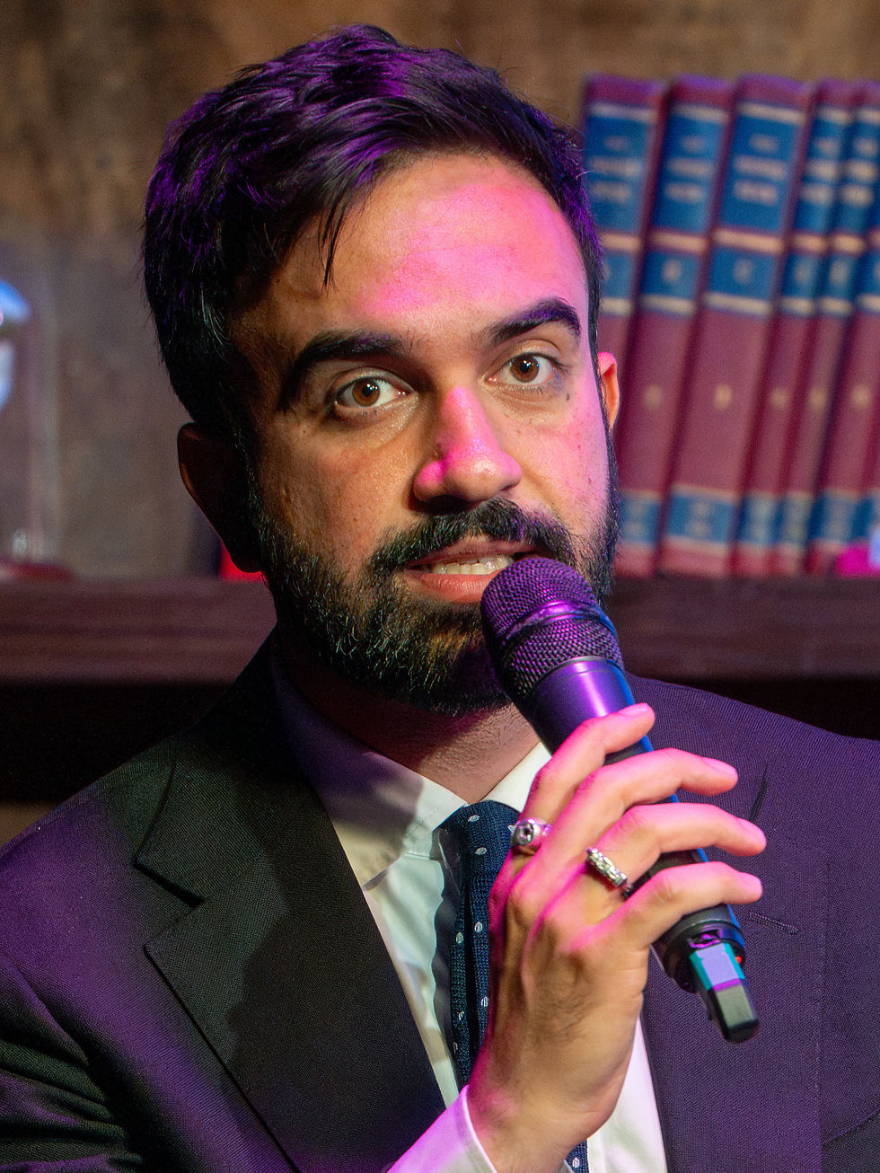 Man with a beard holding a microphone, wearing a suit, in front of a bookshelf. The setting is warmly lit, with a focused expression.