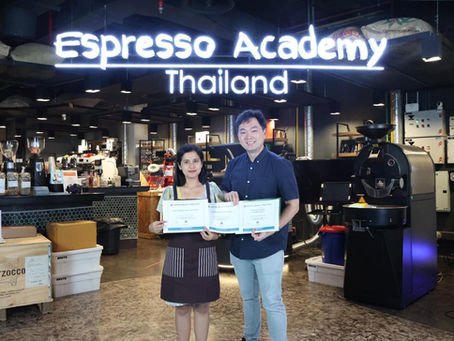 Two people hold coffee-education documents beneath an Espresso Academy Thailand sign in a training space.