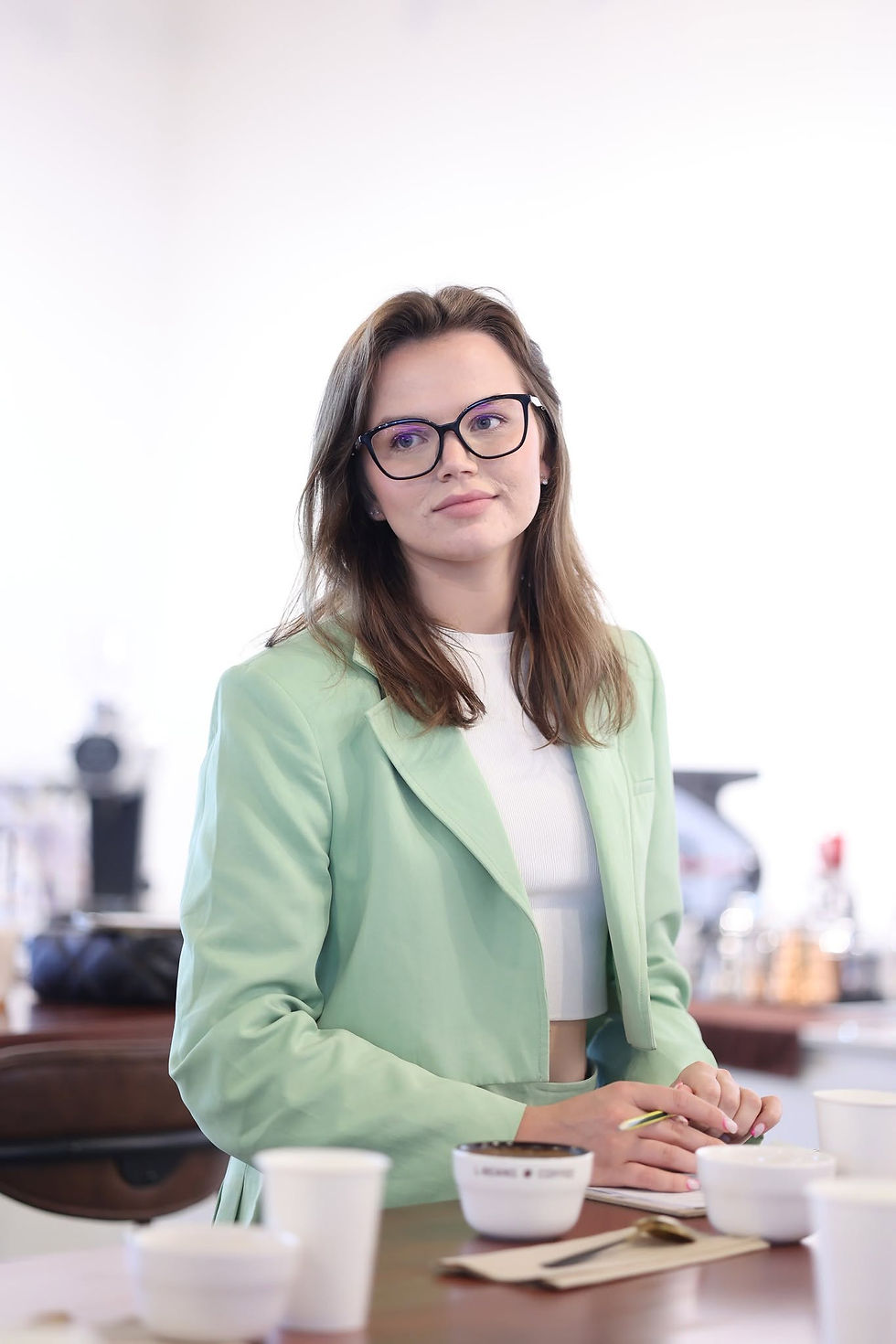 A participant in a pale green jacket sits at a cupping table with coffee cups, a spoon and notes.