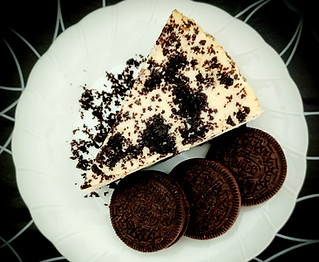 Stewarts' Cheesecakes Cookies and Cream cheesecake slice on a white plate with 3 oreos to the side and oreo crumbs on the plate. The plate is on a black table cloth with white swirls