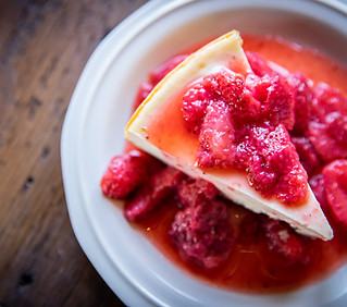 Stewarts' Cheesecakes Classic cheesecake slice on a white plate with strawberries in syrup on the top of the slice. The plate is on a wooden table and is offset to the right