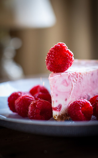 White chocolate raspberry cheesecake slice on a blue grey plate with raspberries around it on the plate and one raspberry on top near the tip of the cheesecake.