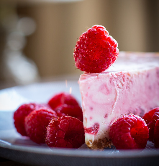 White chocolate raspberry cheesecake slice on a blue grey plate with raspberries around it on the plate and one raspberry on top near the tip of the cheesecake.