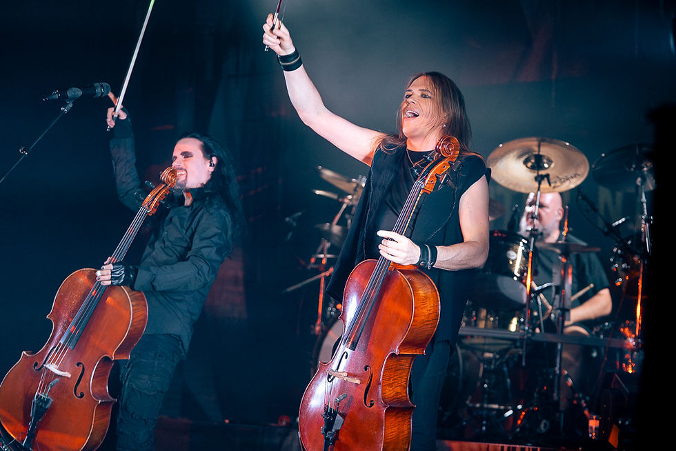 Musicians with cellos perform energetically on stage, with a drummer in the background. Dim lighting, vibrant mood, black and orange attire.