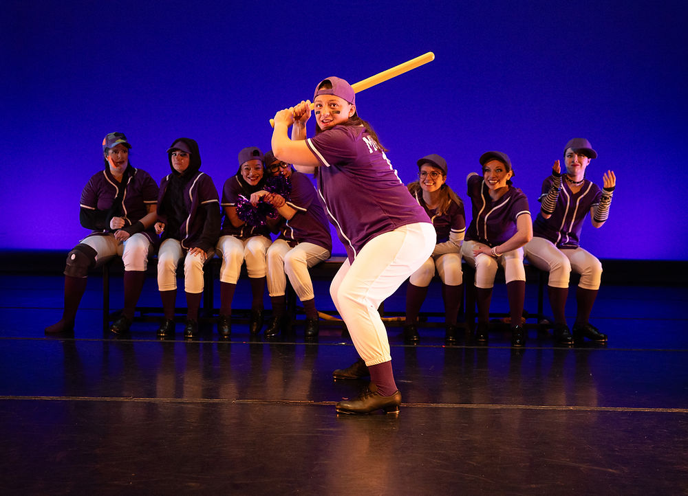 A dancer in full baseball uniform stands ready to swing a bat, a group of 8 players sit on a bench behind her with various expressions.