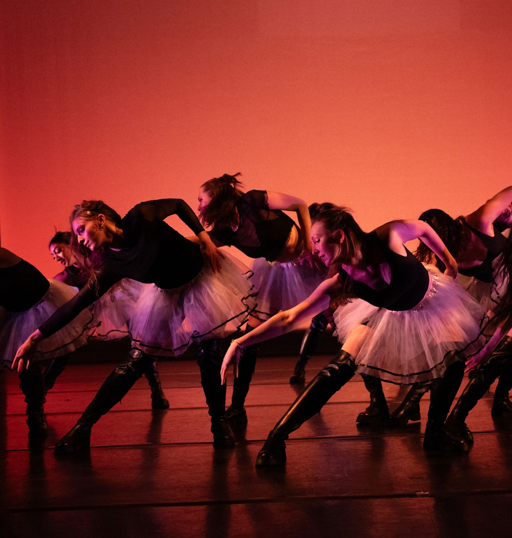 A group of dancers wearing black tops and white tutus, leaning and reaching down towards the ground