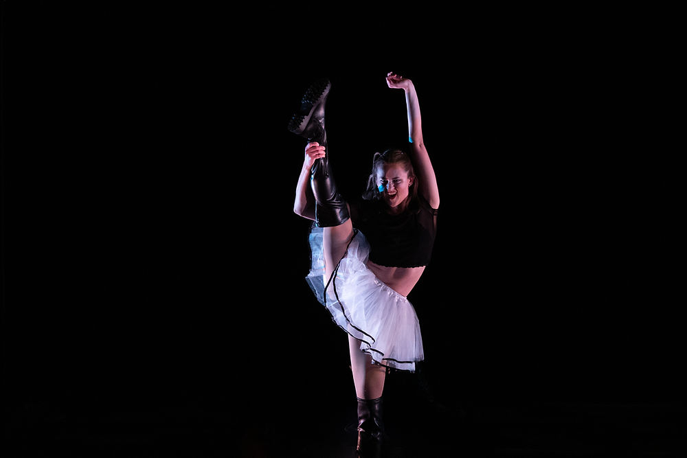 Dancer in black top and white tutu, lifts leg towards head in flexible extension.