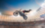 mostra um momento eletrizante de motocross freestyle dentro de um estádio lotado. O piloto, em pleno voo, executa uma manobra radical conhecida como "superman", onde seu corpo está completamente esticado para trás, com os pés soltos da moto e os braços segurando o guidão — como se estivesse voando. O cenário é um espetáculo à parte: o estádio está iluminado por potentes refletores, com uma multidão assistindo e vibrando ao fundo. O céu ao entardecer adiciona um clima épico à cena, enquanto um telão exibe a performance, reforçando a grandiosidade do evento. Essa imagem captura a emoção, ousadia e habilidade extrema que tornam o motocross freestyle um verdadeiro show de adrenalina sobre duas rodas.