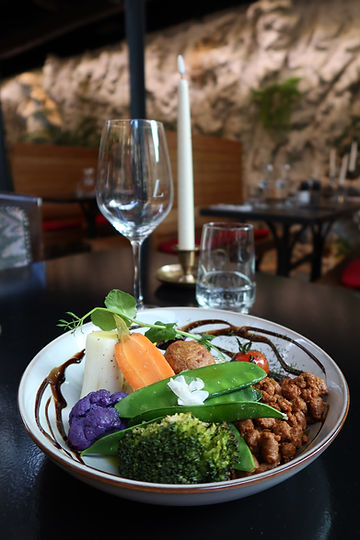 Plat de restaurant végétarien : bol de légumes colorés (chou-fleur violet, brocoli, poireau, haricot mange-tout, tomate cerise, courgette jaune) sur lit de houmous, accompagnés de boulette végétale et haché végétal sur une table dressée