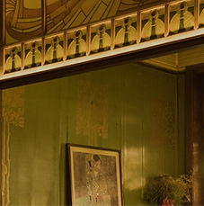 Intérieur de style Art Nouveau montrant un mur vert décoré de motifs d'arbres stylisés dorés. Au-dessus, une frise architecturale complexe avec des formes géométriques et des vitraux dans des tons ambrés