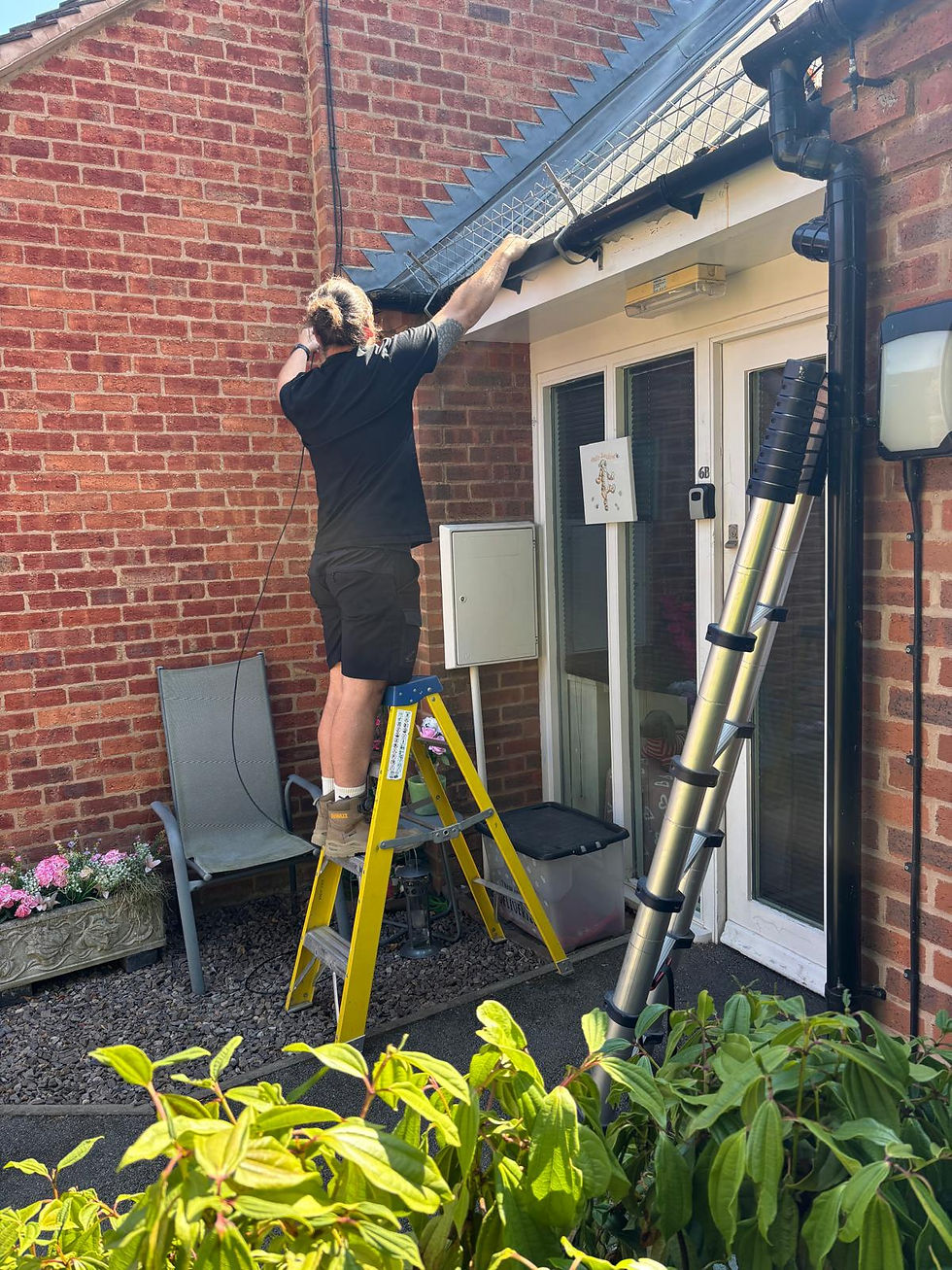 Midland Alarm Services East engineer on a step ladder fitting a CCTV camera under the roof eaves of a customer's home, with cable routed down the brickwork near the back door.