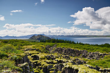 A view across moss-covered lava rocks and a deep crevice toward the large lake Þingvallavatn in Iceland's Þingvellir National Park under a blue sky.