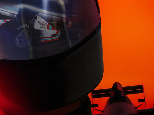 A dramatic close-up of a racing helmet with a reflective visor showing an open-wheel race car. Behind the helmet, a silhouetted Formula-style car is seen against a vivid gradient sunset of red and orange. The overall composition is stylized and intense, evoking high-speed racing and cinematic tension.