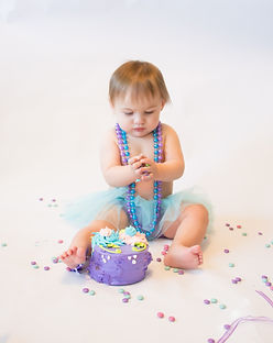 One year old smashing and eating a birthday cake wearing bead necklace on a white backdrop.