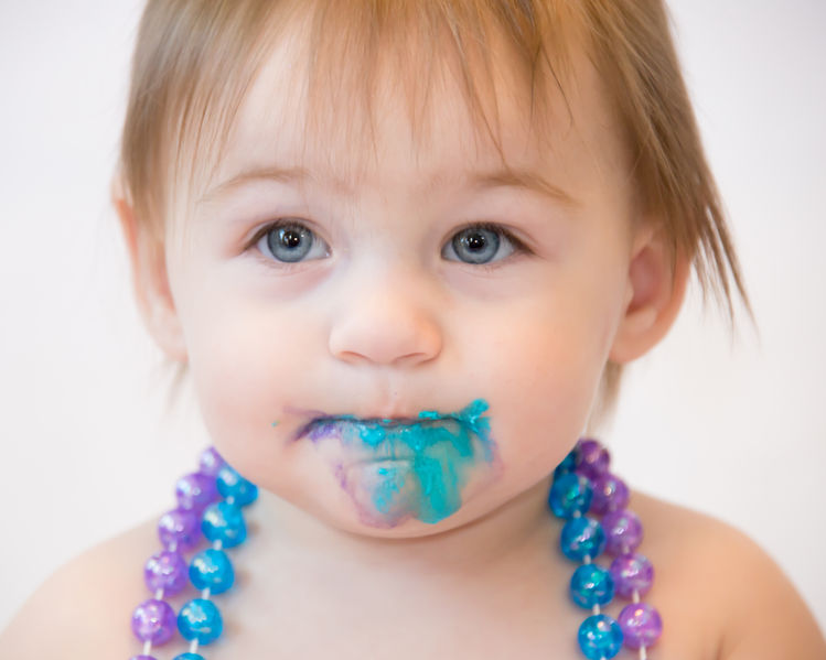 1 year old girl cake smash portrait on white backdrop with small purple cake and matching beads
