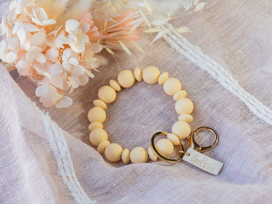 Pale yellow beaded wristlet keychain with gold hardware, styled on soft fabric with florals.