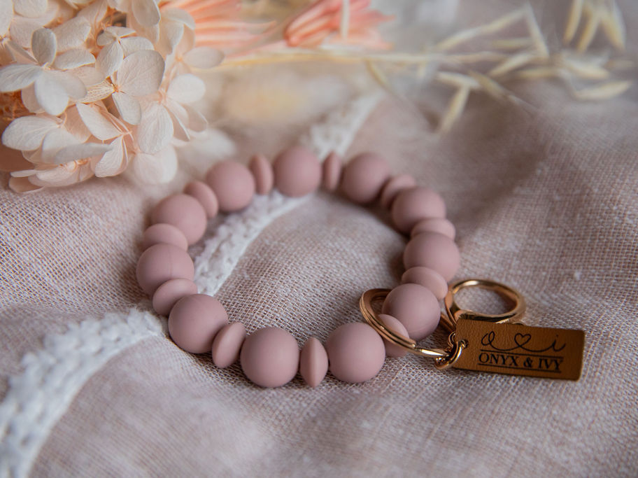Blush beaded wristlet keychain with gold hardware, styled on soft fabric with florals.