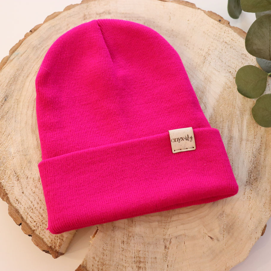 Fuchsia knit toque with Onyx & Ivy logo tag, styled on wood round with greenery.
