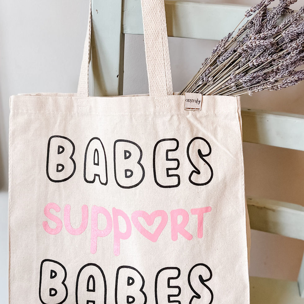 Canvas tote bag with “Babes Support Babes” design in black and pink, styled with lavender stems against a chair.