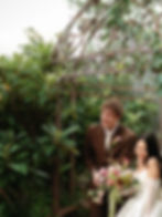 A bride with a pink and lime green bouquet laughs with her groom in a brown custom suite under an ir