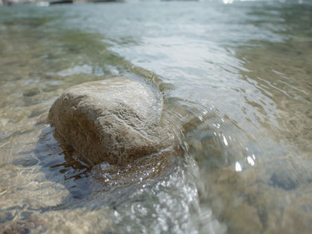 Clear river with smooth rocks, gentle water flow around a prominent stone. Green foliage and rocky banks in the background. Tranquil scene. Symbolizing how gentle movement can be better than hard training.