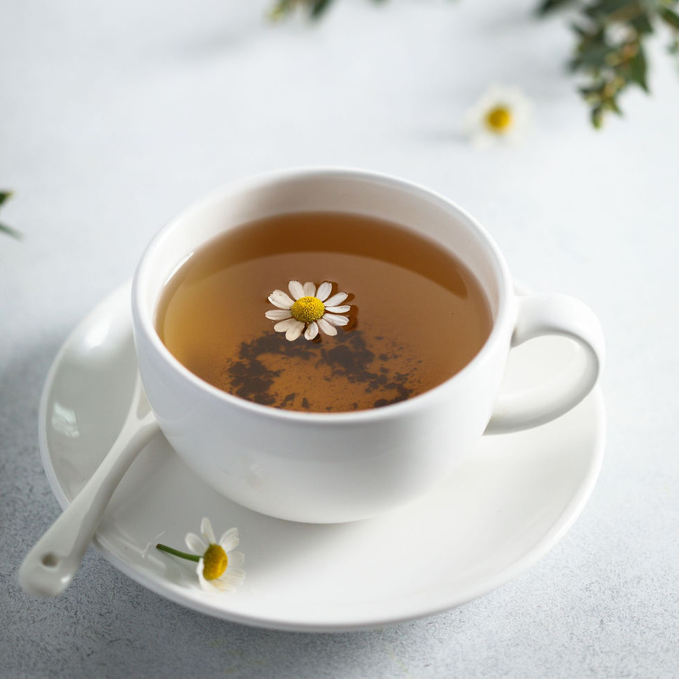 White cup with chamomile tea, a daisy floating on top. Cup on saucer with a spoon. Soft, light background with greenery for a calming mood.