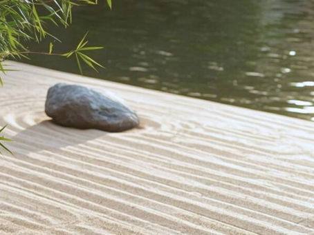Zen garden with raked sand patterns, a stone, and bamboo in the foreground. Calm water reflects sunlight in the background. Symbolizing importance of rest.