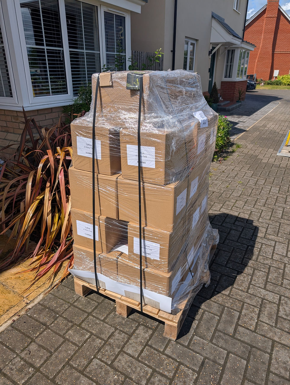 Pallet of shrink-wrapped cardboard boxes labeled Suffolk A Story Told through food, drink and people on a sunny driveway beside a house.