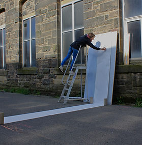 A large piece of paper is attached to a wall and trailing on the floor. A woman is stood up a ladder reaching over to it. it looks like she could be drawing.