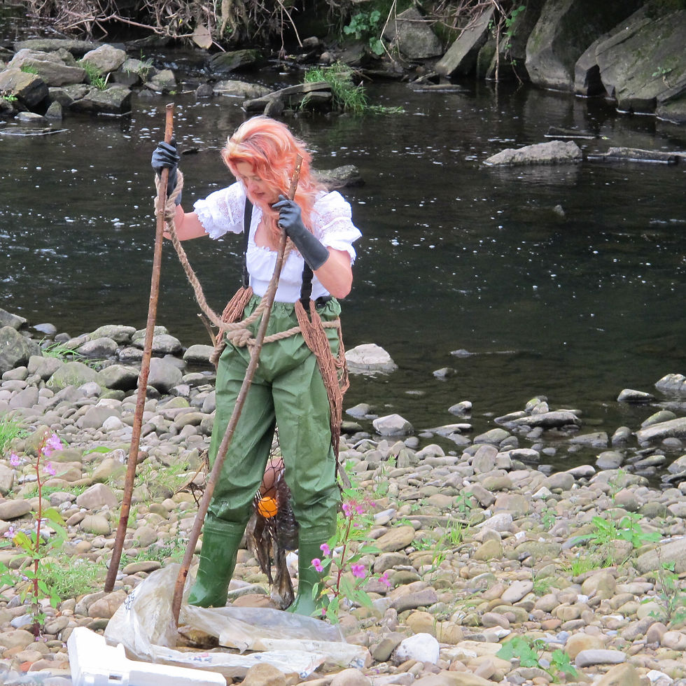 A woman stands at the edge of a river bank holding two sticks and covered in detritus