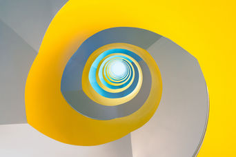An abstract look-up study of a spiral staircase, emphasizing the clean geometry and striking color contrast between vibrant yellow, concrete gray, and a central blue gradient. 
