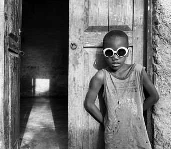 A boy stands wearing sunglasses next to an open door flooded with light.