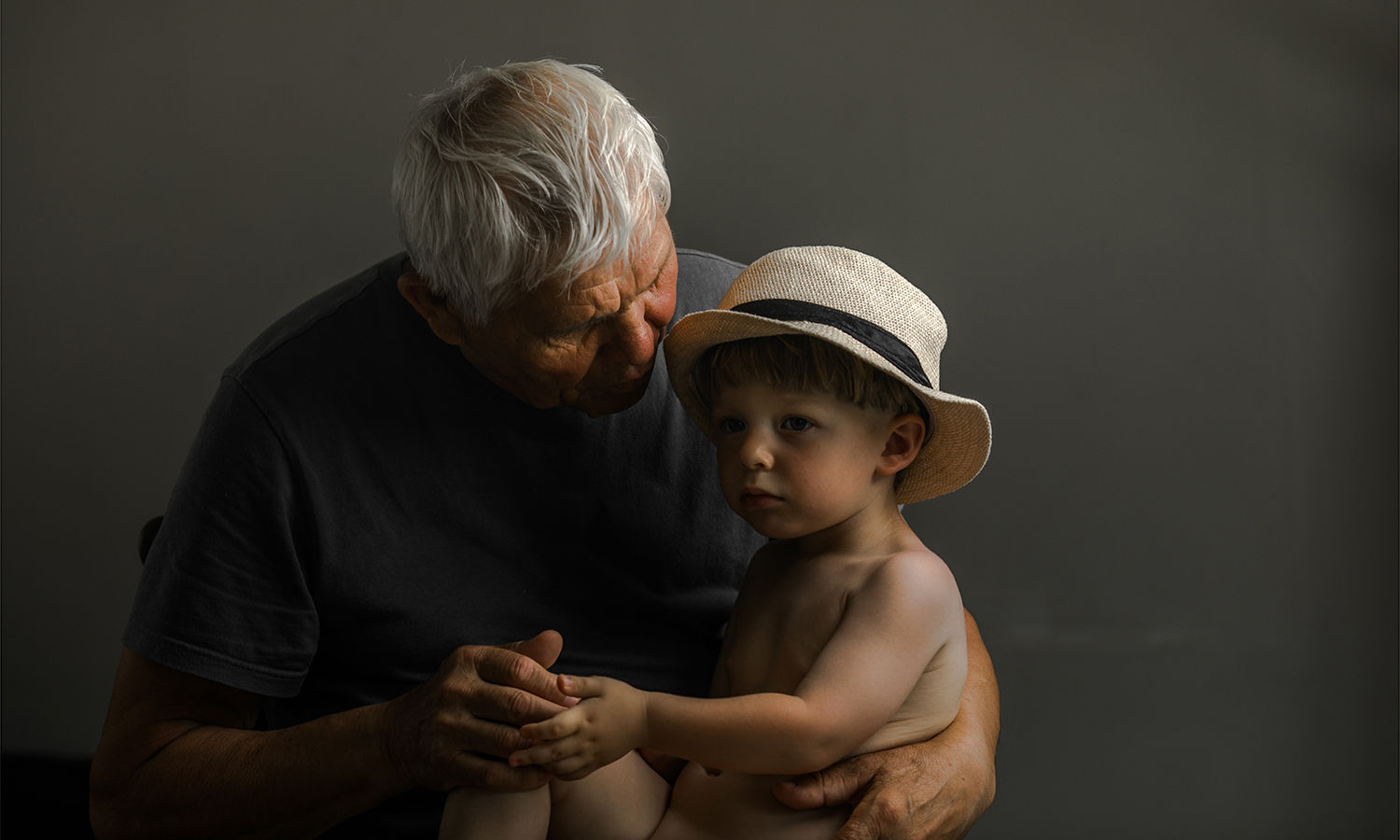 This portrait captures a quiet, profound moment between an elderly man and a young child, embodying the passage of time and the tender fragility of life. The grandfather’s weathered hands gently hold the child, his face leaning in with a soft, protective presence.