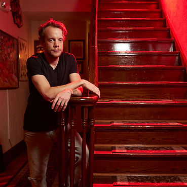 Man in black shirt leaning on dramatic red-lit wooden staircase.