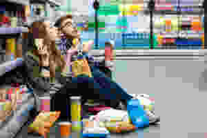 A male and female sitting in a grocery store aisle eating and drinking snacks.