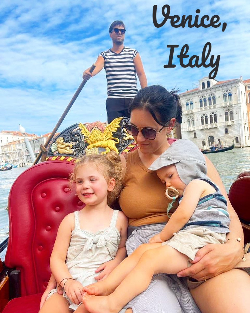 Woman with two kids on a gondola in Venice, Italy. Gondolier rowing in background. Text reads "Venice, Italy." Sky is blue and clear.