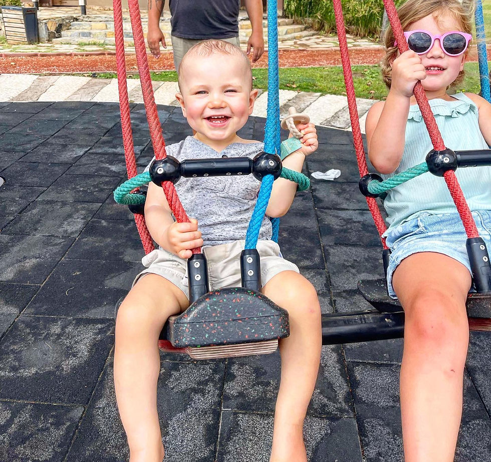Two kids swing joyfully on colorful swings in a playground, smiling and wearing casual summer clothes. Background features greenery.