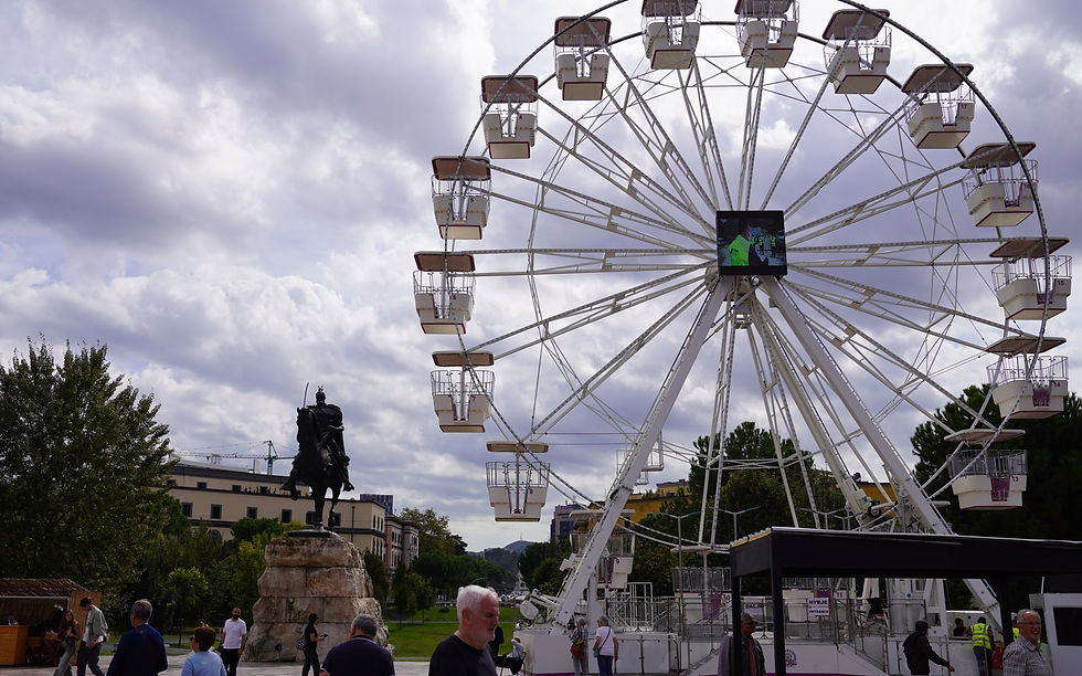 People walking in a plaza with a large ferris wheel and an equestrian statue. Cloudy sky, trees, and buildings in the background create a lively scene.