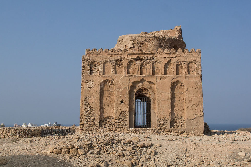Ancient stone structure with arched doorway, against a clear blue sky. Rocky terrain surrounds, with distant sea visible in the background.