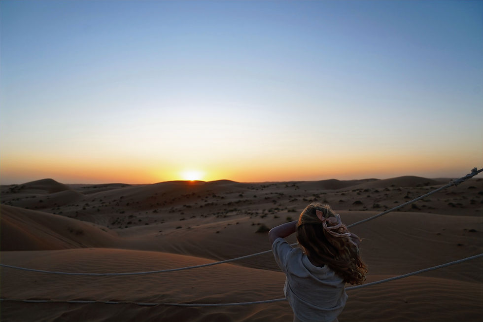 A person with a pink bow watches a serene desert sunset. Sand dunes stretch into the distance under a gradient sky, evoking tranquility.