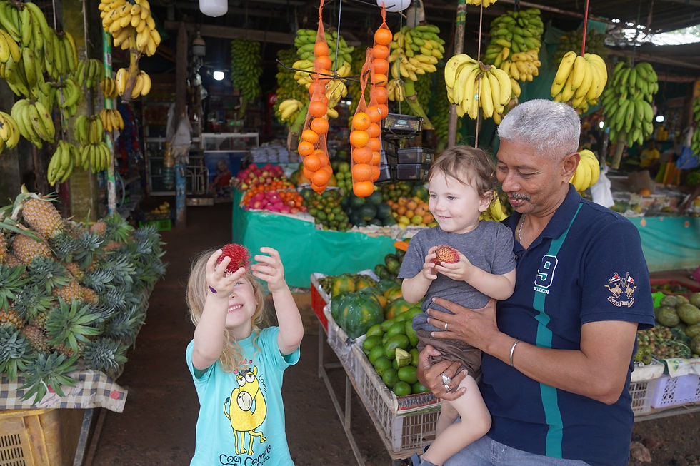 Two kids hold red fruit joyfully at a vibrant market stand with heaps of bananas and oranges. An older man smiles warmly beside them.