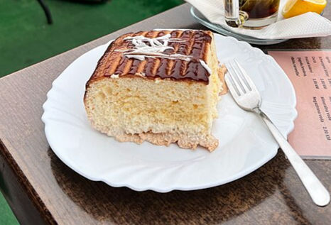 Slice of layered cake on a white plate with a fork beside it, set on a wooden table. Background shows a menu and a glass with lemon.