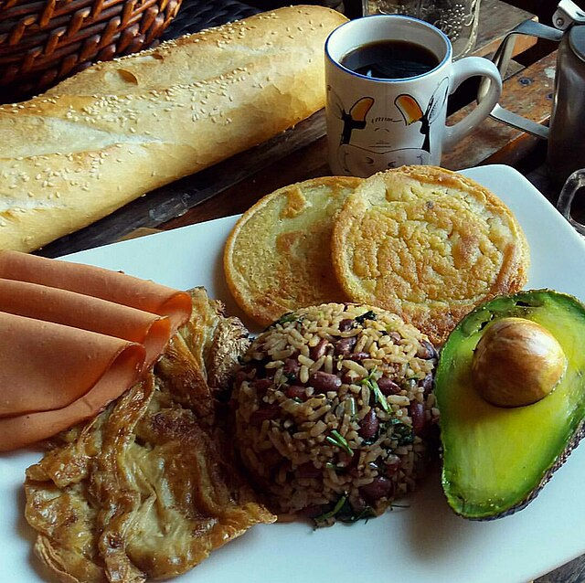 Breakfast plate with rice, beans, plantain, avocado, arepas, ham, egg, baguette, and a toucan mug of coffee on a wooden table.