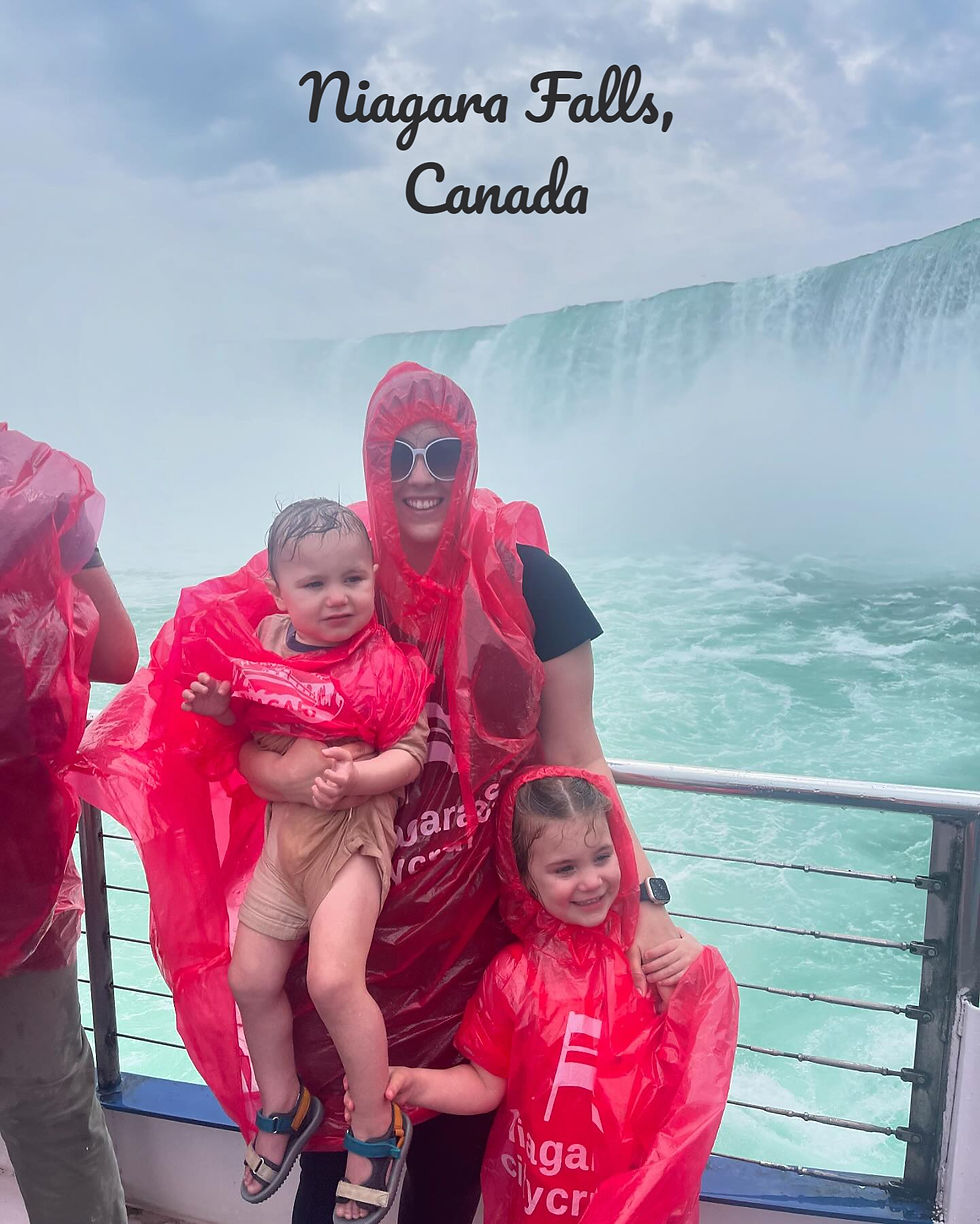 People in red ponchos on a boat at Niagara Falls. A smiling adult holds a child; another child stands nearby. Text: Niagara Falls, Canada. Waterfall mist in background.