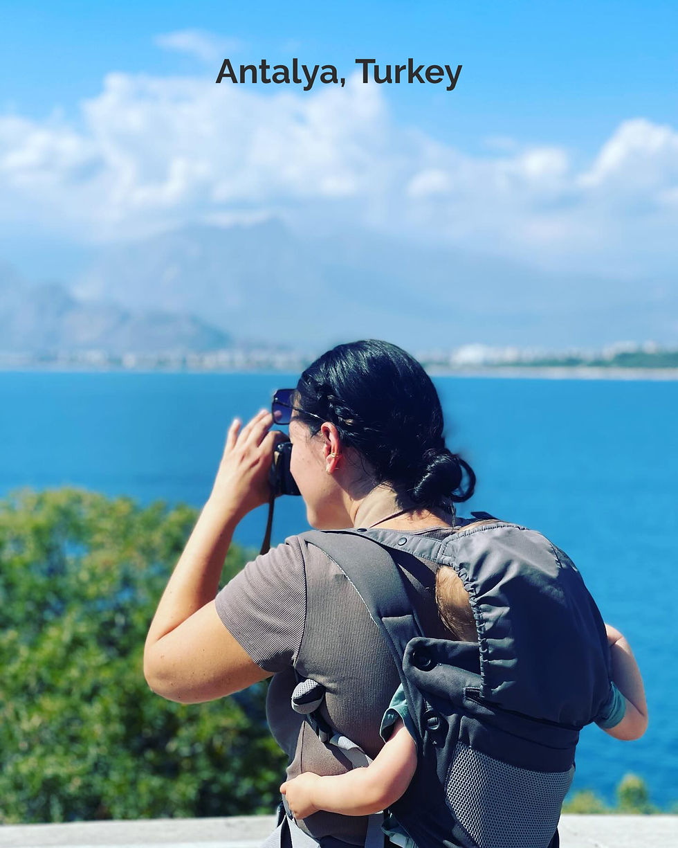 Woman with baby in carrier takes photos by blue sea in Antalya, Turkey. Greenery and mountains in the background, clear skies.