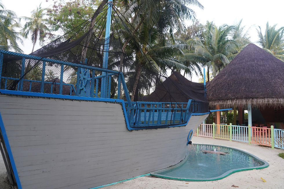 Playground with a large, blue and gray pirate ship structure, palm trees, a hut, and colorful fence in the background. Relaxed atmosphere.