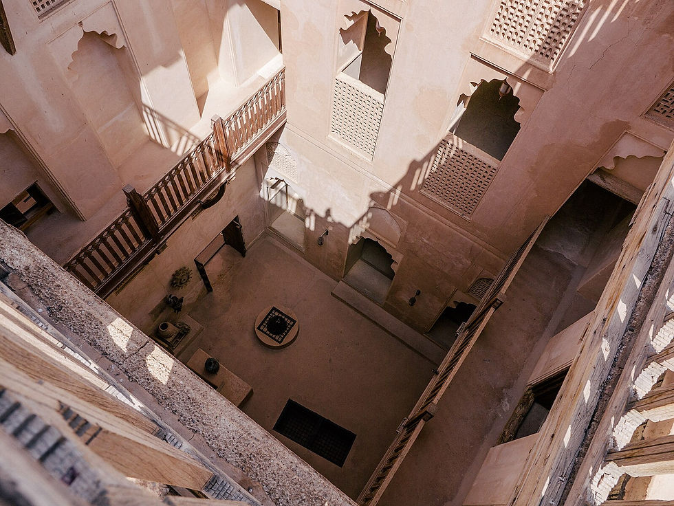 Courtyard view of historic beige building with wooden railings and decorative windows. Shadows cast intricate patterns on walls and stairs.